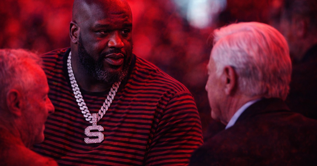 Towering bald man with jeweled pendant speaking candidly in crimson-lit arena crowd