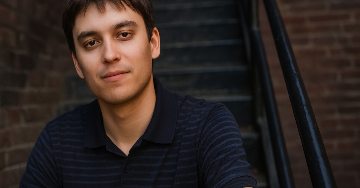 Young man in navy polo shirt leaning against weathered red brick wall, side angle portrait
