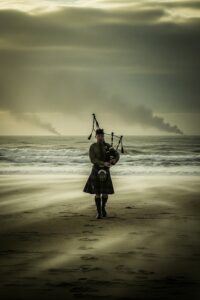 Lone Scottish piper in kilt playing bagpipes on a stormy WWII beach at dawn