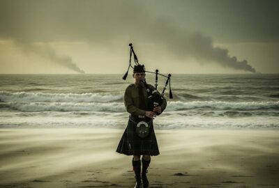 Lone Scottish piper in kilt playing bagpipes on a stormy WWII beach at dawn
