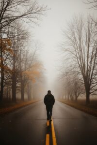 Lone man walking a foggy empty road at dusk, silhouette fading into mist