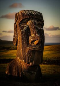 A solitary Easter Island moai statue glowing in dusk side-light, with windswept volcanic grassland and the Pacific Ocean behind.