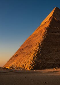 The Great Pyramid of Khufu rising against a deep blue Egyptian sky at golden hour, weathered limestone glowing warm amber