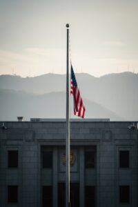 An American flag rising up a tall flagpole outside a stone embassy building under a pale sky