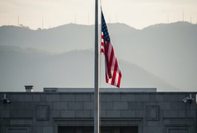 An American flag rising up a tall flagpole outside a stone embassy building under a pale sky