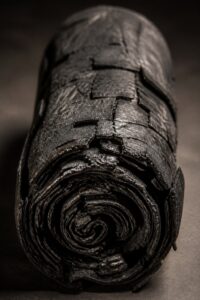 A blackened, carbonised Herculaneum papyrus scroll fused solid, lit dramatically against a dark background