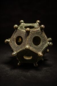 A hollow twelve-sided Roman bronze dodecahedron with round holes of different sizes and knobbed corners, lit dramatically on a dark background