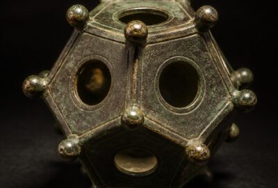 A hollow twelve-sided Roman bronze dodecahedron with round holes of different sizes and knobbed corners, lit dramatically on a dark background