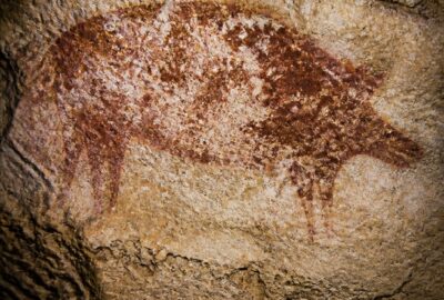 The world's oldest story cave painting: a faded red-ochre scene of a Sulawesi warty pig and human-like figures on a limestone cave ceiling at Leang Karampuang