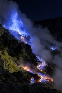 Electric-blue flames of burning sulfur gas rising from vents in the dark crater of Kawah Ijen volcano, East Java, with molten sulfur glowing on black rock