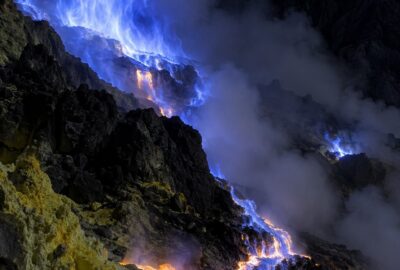 Electric-blue flames of burning sulfur gas rising from vents in the dark crater of Kawah Ijen volcano, East Java, with molten sulfur glowing on black rock