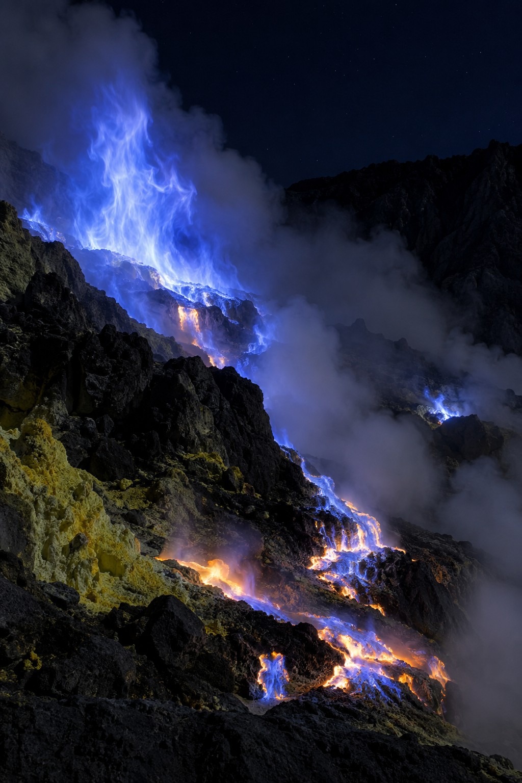 Electric-blue flames of burning sulfur gas rising from vents in the dark crater of Kawah Ijen volcano, East Java, with molten sulfur glowing on black rock
